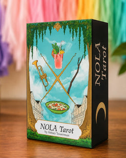A boxed tarot card deck standing on a wooden surface. The front of the box shows an illustrated scene with a blue sky, green vines, crossed gold swords, a pink drink, a bowl of food, and a black side panel with the title 'NOLA Tarot' and the author name 'by Ashley Testerman