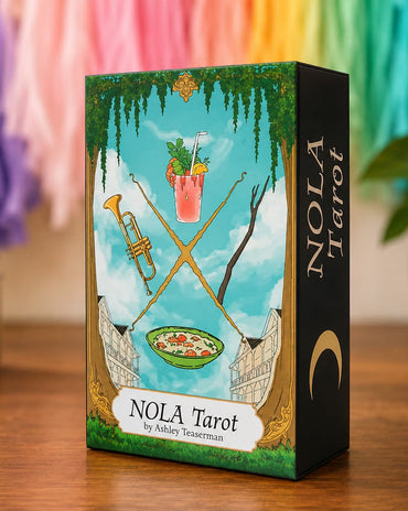 A boxed tarot card deck standing on a wooden surface. The front of the box shows an illustrated scene with a blue sky, green vines, crossed gold swords, a pink drink, a bowl of food, and a black side panel with the title 'NOLA Tarot' and the author name 'by Ashley Testerman