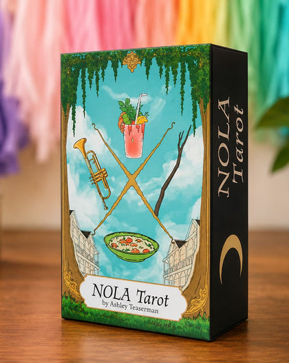 A boxed tarot card deck standing on a wooden surface. The front of the box shows an illustrated scene with a blue sky, green vines, crossed gold swords, a pink drink, a bowl of food, and a black side panel with the title 'NOLA Tarot' and the author name 'by Ashley Testerman