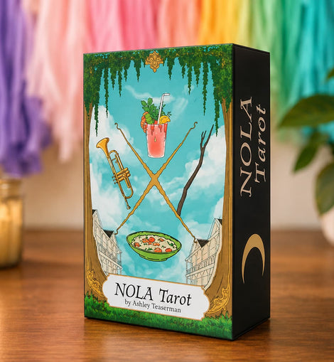 A boxed tarot card deck standing on a wooden surface. The front of the box shows an illustrated scene with a blue sky, green vines, crossed gold swords, a pink drink, a bowl of food, and a black side panel with the title 'NOLA Tarot' and the author name 'by Ashley Testerman