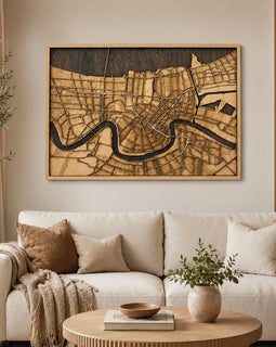 A large framed wooden map wall art piece depicting a winding river through a city, displayed above a cream sofa in a styled living room setting.