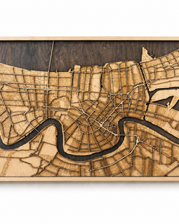 A rectangular wooden city map wall art piece featuring a laser-engraved street grid and a winding river, shown in a light wood frame against a plain white background.
