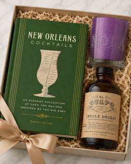 A gift box set containing a green 'New Orleans Cocktails' book by Sarah Bairo, a bottle of El Cuafo Creole Orgat liqueur, and a purple candle, all nestled in crinkle paper and tied with a beige ribbon on a marble surface.