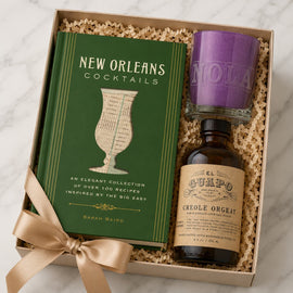 A gift box set containing a green 'New Orleans Cocktails' book by Sarah Bairo, a bottle of El Cuafo Creole Orgat liqueur, and a purple candle, all nestled in crinkle paper and tied with a beige ribbon on a marble surface.