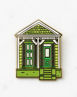 A small enamel pin shaped like a house, featuring green shutters and doors, a green shingled roof, white trim, and a gold-tone metal outline, shown on a light marble-like background.