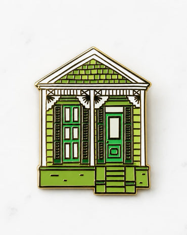 A small enamel pin shaped like a house, featuring green shutters and doors, a green shingled roof, white trim, and a gold-tone metal outline, shown on a light marble-like background.