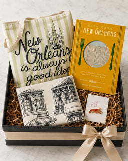 A New Orleans-themed gift basket in a black box with a cream ribbon, containing a striped tote bag with 'New Orleans is always good idea' text, a yellow book titled 'City Eats New Orleans' by Beth Sandowich, a small printed pillow with architectural sketches, and a small card.
