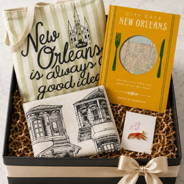 A New Orleans-themed gift basket in a black box with a cream ribbon, containing a striped tote bag with 'New Orleans is always good idea' text, a yellow book titled 'City Eats New Orleans' by Beth Sandowich, a small printed pillow with architectural sketches, and a small card.