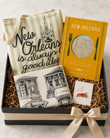 A New Orleans-themed gift basket in a black box with a cream ribbon, containing a striped tote bag with 'New Orleans is always good idea' text, a yellow book titled 'City Eats New Orleans' by Beth Sandowich, a small printed pillow with architectural sketches, and a small card.