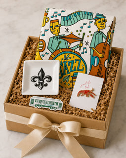 A gift box filled with curated items on a light marble surface: a kraft gift box with a cream ribbon, a patterned jazz-themed notebook or card, a small white square dish with a black fleur-de-lis, a small card with a red lobster illustration, and a green-and-white ticket-like accessory.