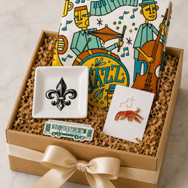 A gift box filled with curated items on a light marble surface: a kraft gift box with a cream ribbon, a patterned jazz-themed notebook or card, a small white square dish with a black fleur-de-lis, a small card with a red lobster illustration, and a green-and-white ticket-like accessory.