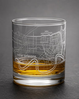 A clear glass rocks tumbler with an etched New Orleans map design, partially filled with amber whiskey, set against a dark gray background.