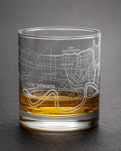 A clear glass rocks tumbler with an etched New Orleans map design, partially filled with amber whiskey, set against a dark gray background.