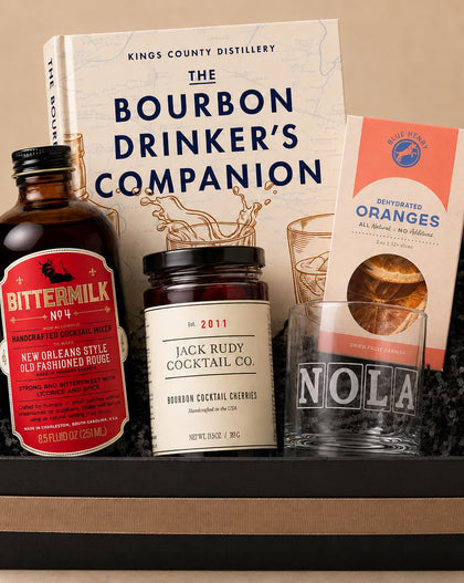 A curated gift basket or whiskey companion set arranged in a black box with a beige band. Visible items include a bottle of Bitter Milk No.4