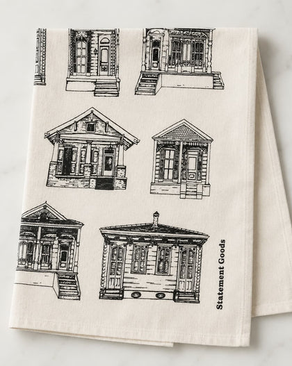 An off-white kitchen tea towel featuring black line-art illustrations of various house frontages, with the words 'Statement Goods' printed vertically on the right side, displayed on a light marble surface.