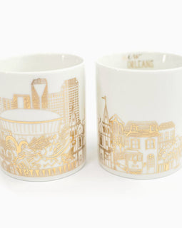 White ceramic New Orleans skyline mug with gold-foil city illustrations and a gold handle, shown from two angles on a white background.