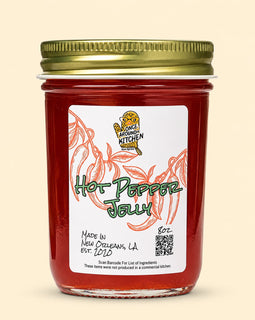 A jar of Fancy Farming Kitchen Hot Pepper Jelly with a gold metal lid and a white label featuring red pepper illustrations, set against a solid light beige background.
