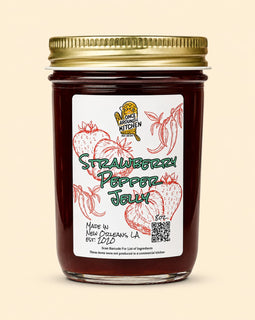 A glass jar of Strawberry Pepper Jelly with a gold metal lid and a white label featuring red strawberry illustrations and green text, set against a solid beige background.