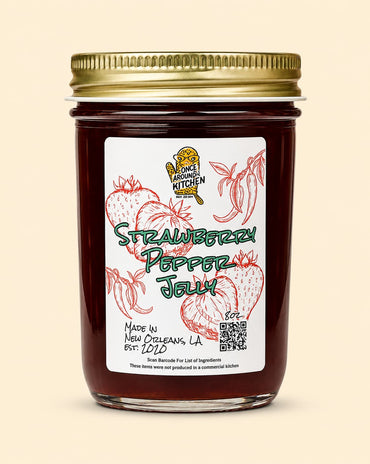 A glass jar of Strawberry Pepper Jelly with a gold metal lid and a white label featuring red strawberry illustrations and green text, set against a solid beige background.