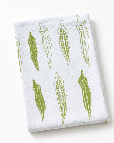 A folded white kitchen towel featuring a green line-art pattern of okra pods, photographed on a clean white background.