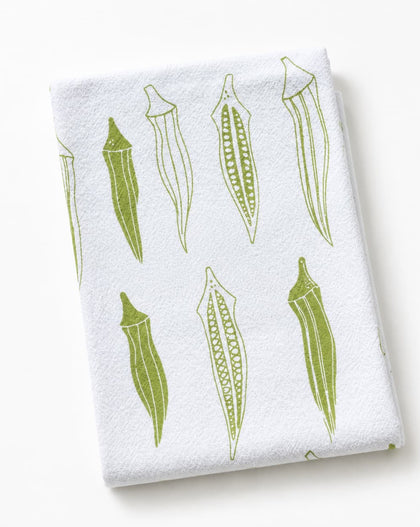 A folded white kitchen towel featuring a green line-art pattern of okra pods, photographed on a clean white background.