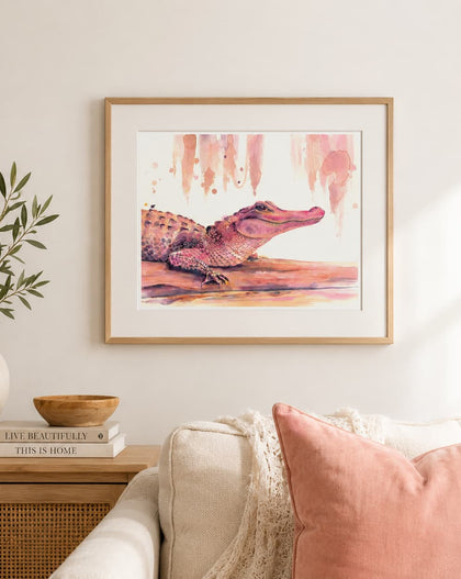 A framed watercolor art print of a pink and brown alligator hanging on a light beige wall above a sofa with a pink pillow, styled in a cozy living room setting.