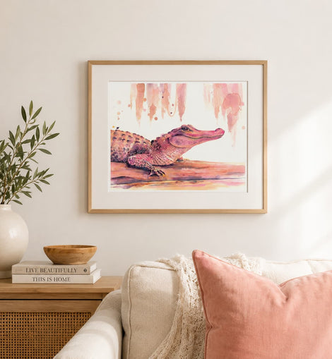 A framed watercolor art print of a pink and brown alligator hanging on a light beige wall above a sofa with a pink pillow, styled in a cozy living room setting.