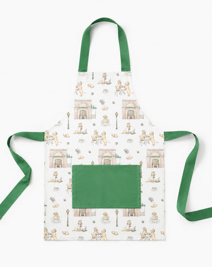 A white apron with a green neck strap and waist ties, featuring a repeating pattern of vintage illustrations of people sitting at tables and a large solid green front pocket.