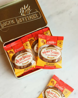 A tin of Aunt Sally's Classic Whole Pralines with several individual packet packs placed in front and one loose packet on a light marble surface. The tin lid is open, showing the brand name 'Aunt Sally's' and a sun emblem.