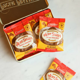 A tin of Aunt Sally's Classic Whole Pralines with several individual packet packs placed in front and one loose packet on a light marble surface. The tin lid is open, showing the brand name 'Aunt Sally's' and a sun emblem.