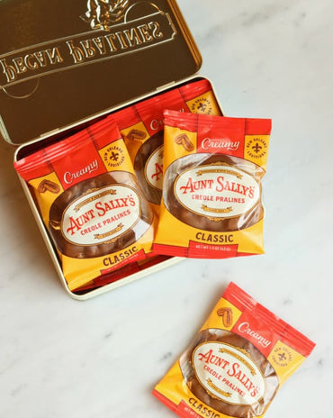 A tin of Aunt Sally's Classic Whole Pralines with several individual packet packs placed in front and one loose packet on a light marble surface. The tin lid is open, showing the brand name 'Aunt Sally's' and a sun emblem.