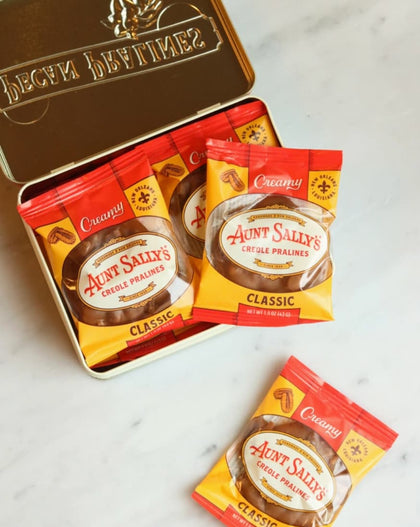 A tin of Aunt Sally's Classic Whole Pralines with several individual packet packs placed in front and one loose packet on a light marble surface. The tin lid is open, showing the brand name 'Aunt Sally's' and a sun emblem.