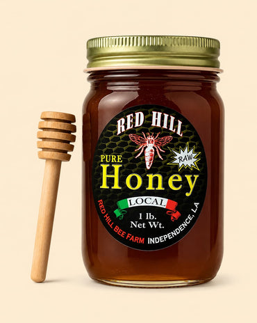 A glass jar of Red Hill Pure Local Honey with a gold metal lid and a wooden honey dipper, set against a solid light beige background.