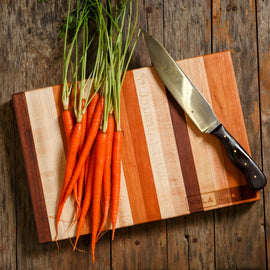 Striped Cherry, Maple, and Walnut cutting board with carrots and a chef’s knife on a rustic wooden table.