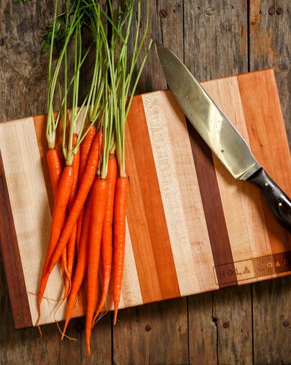 Striped Cherry, Maple, and Walnut cutting board with carrots and a chef’s knife on a rustic wooden table.