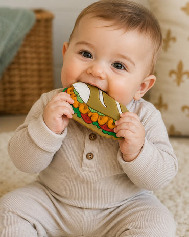A baby wearing a light beige ribbed long-sleeve romper sits on a cream textured rug, holding a colorful fabric teething toy shaped like a shrimp po boy