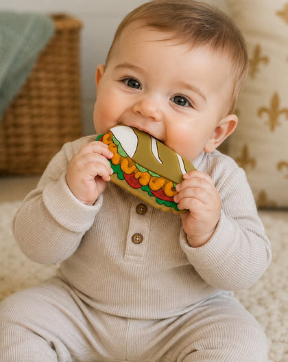 A baby wearing a light beige ribbed long-sleeve romper sits on a cream textured rug, holding a colorful fabric teething toy shaped like a shrimp po boy