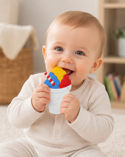 A baby sitting on a carpeted floor holding and biting a colorful teething toy shaped like a snowball in a softly lit indoor room.