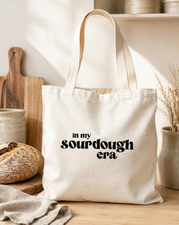 A cream canvas tote bag with black text reading 'in my sourdough era' sits on a wooden table in a warm, rustic kitchen setting with a loaf of bread, cutting board, and ceramic dishes in the background.
