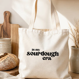 A cream canvas tote bag with black text reading 'in my sourdough era' sits on a wooden table in a warm, rustic kitchen setting with a loaf of bread, cutting board, and ceramic dishes in the background.