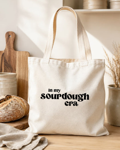 A cream canvas tote bag with black text reading 'in my sourdough era' sits on a wooden table in a warm, rustic kitchen setting with a loaf of bread, cutting board, and ceramic dishes in the background.