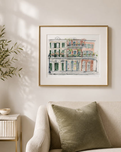 A framed watercolor art print of a classic French or European building facade with green shutters and warm terracotta tones, hanging on a beige wall above a sofa in a styled living room.
