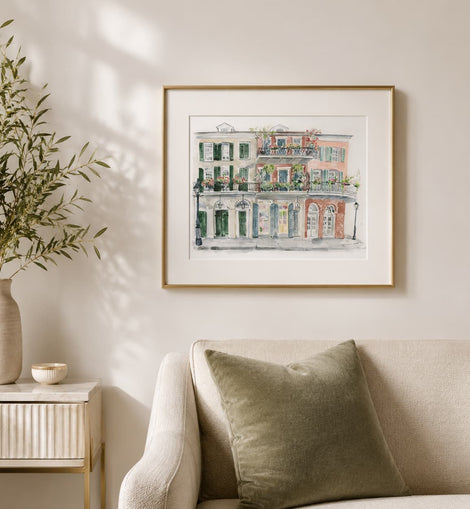 A framed watercolor art print of a classic French or European building facade with green shutters and warm terracotta tones, hanging on a beige wall above a sofa in a styled living room.