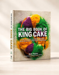 The Big Book of King Cake cookbook by Matt Haines, standing upright on a light wooden surface against a warm beige background. The front cover features a colorful Mardi Gras-themed cake illustration with green, purple, and orange tones on a textured light beige background.