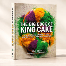 The Big Book of King Cake cookbook by Matt Haines, standing upright on a light wooden surface against a warm beige background. The front cover features a colorful Mardi Gras-themed cake illustration with green, purple, and orange tones on a textured light beige background.