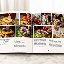 An open cookbook spread displayed on a light marble surface against a white background. The pages feature multiple color food photographs, a quote on the left page, and two columns of text.