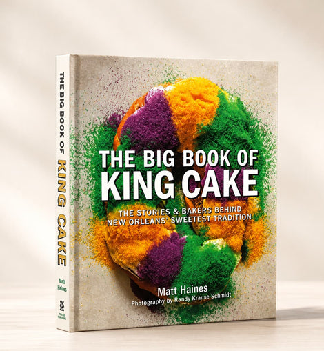 The Big Book of King Cake cookbook by Matt Haines, standing upright on a light wooden surface against a warm beige background. The front cover features a colorful Mardi Gras-themed cake illustration with green, purple, and orange tones on a textured light beige background.