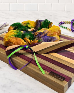 A striped wooden cutting board with purple and light wood bands, tied with a green and purple ribbon, displayed on a white marble countertop with Mardi Gras-themed bread and beads in the background.