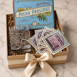 A gift box set featuring Aunt Sally's Pecan Pralines, including a blue branded tin, a clear glass etched with a map outline, and four square coasters printed with illustrations of local buildings, all arranged in a kraft box with shredded filler and a cream ribbon on a marble surface.