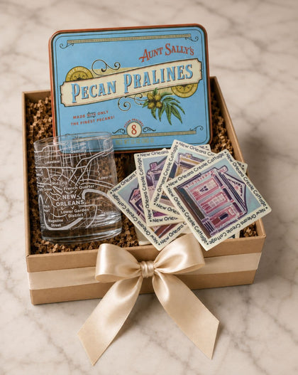 A gift box set featuring Aunt Sally's Pecan Pralines, including a blue branded tin, a clear glass etched with a map outline, and four square coasters printed with illustrations of local buildings, all arranged in a kraft box with shredded filler and a cream ribbon on a marble surface.